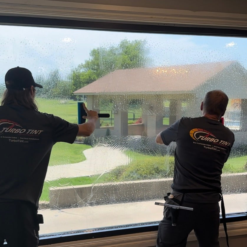 Two workers from Turbo Tint applying window tinting to a large glass window at a community park 