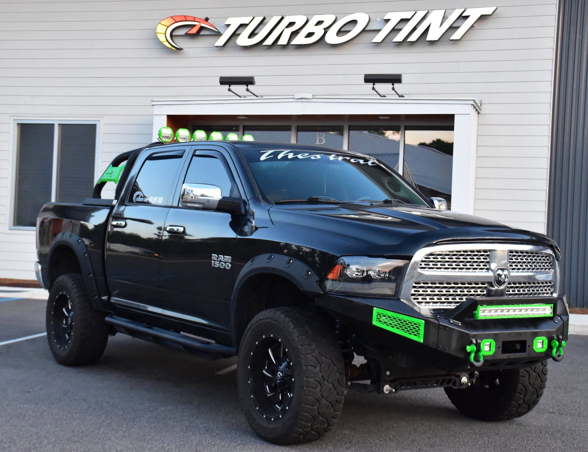 A black Ram 1500 pickup truck parked in front of a building with a large 'TURBO TINT' sign. The truck is equipped with a custom front bumper, green accent grilles, and green LED light bars, with a custom white script decal on the windshield.